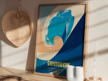 A framed surf poster with abstract blue and beige wave design, titled Cloudbreak Tavarua Island Fiji, is displayed on a wooden surface next to a woven fan and decorative candle.