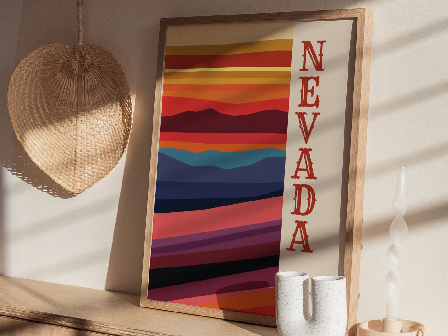A framed Nevada poster with colorful desert landscape stripes and bold red lettering is displayed on a wooden surface, accompanied by a woven wall decor and a white candle.