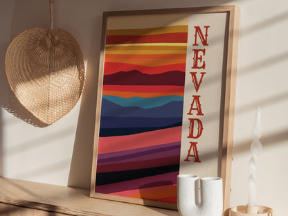 A framed Nevada poster with colorful desert landscape stripes and bold red lettering is displayed on a wooden surface, accompanied by a woven wall decor and a white candle.
