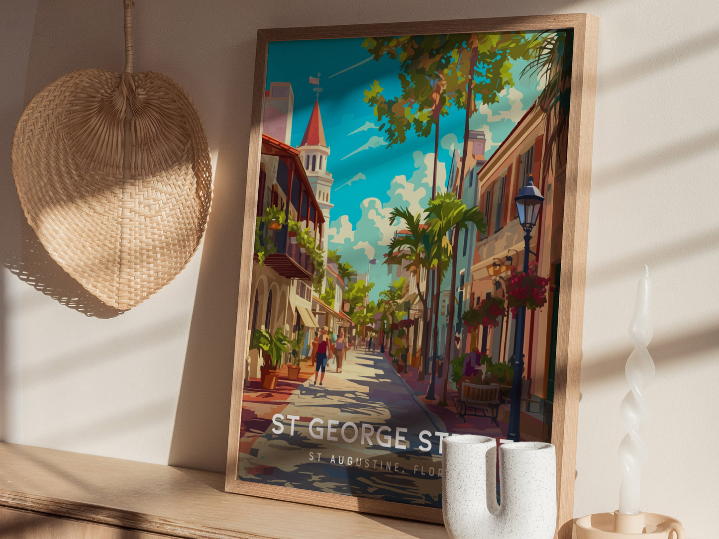 A framed poster of St. George Street in St. Augustine, Florida, features vibrant colors and a picturesque street scene. It rests on a wooden shelf beside a woven fan and.