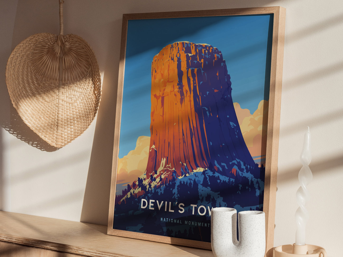 A framed poster of Devils Tower with vibrant colors is displayed on a wooden shelf, accompanied by a woven wall decoration and a twisted candle.