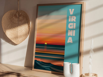 Framed poster with VIRGINIA text, featuring a vibrant sunset over an abstract ocean. It rests on a wooden surface with a woven wall decoration and white ceramic items nearby.