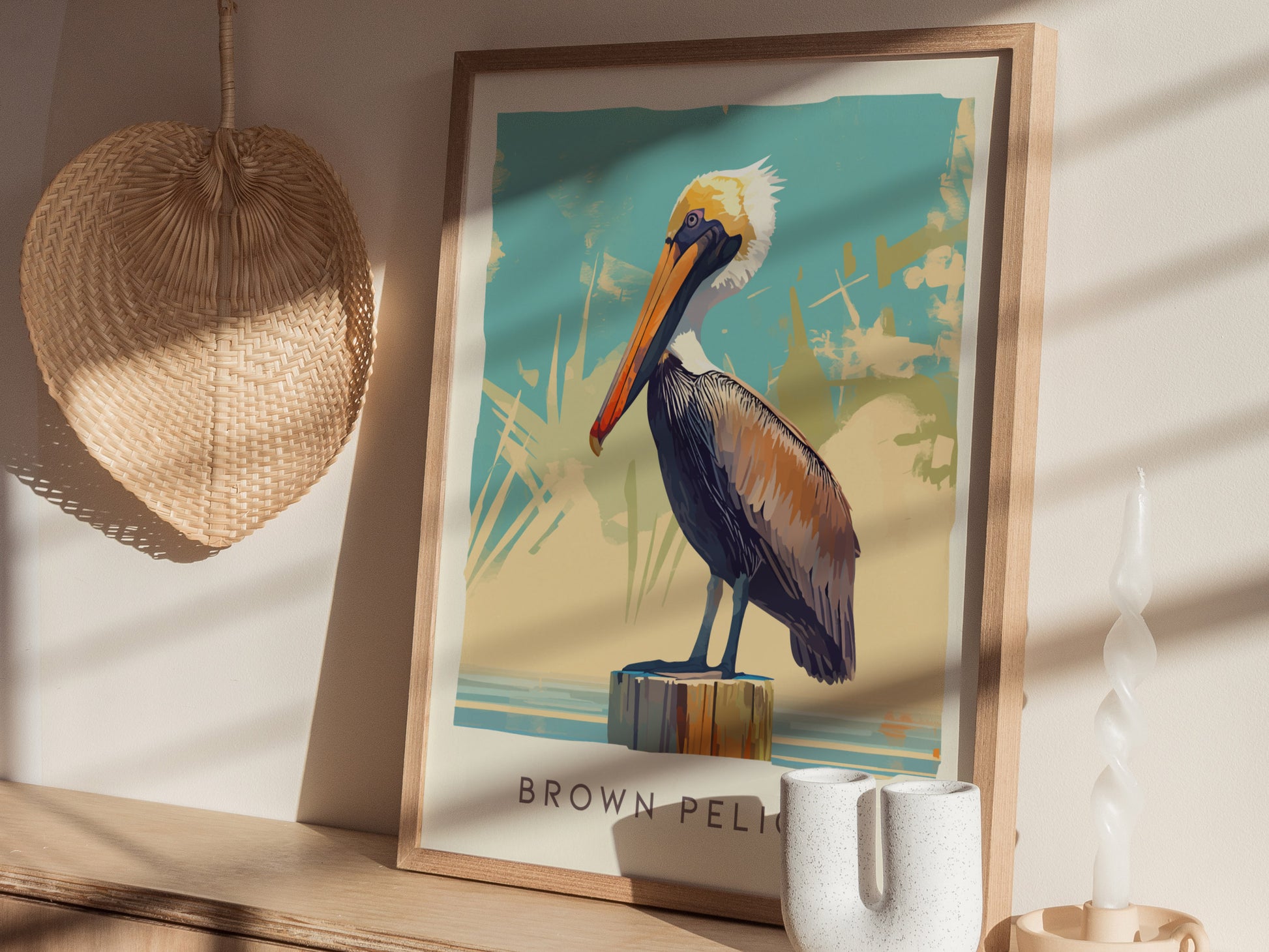 A framed poster of a brown pelican perched on a post, displayed on a shelf with a woven wall decoration and a twisted white candle nearby.