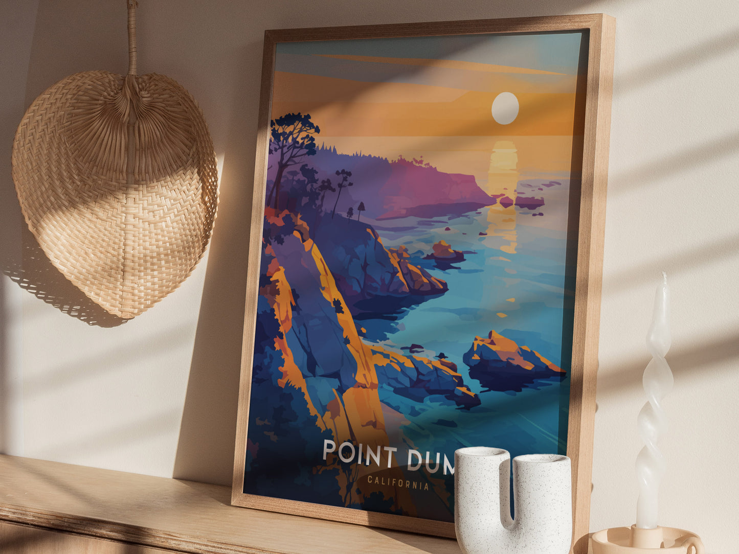 A framed Point Dume poster depicting cliffs and ocean under a setting sun rests on a wooden shelf. Nearby are a woven wall decoration and a unique white candle.