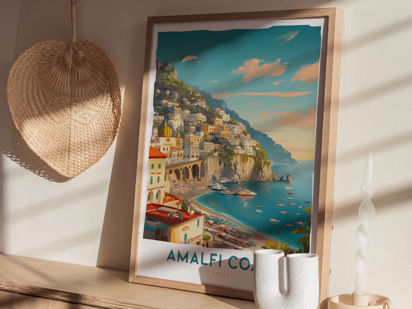 A framed Amalfi Coast travel print rests on a wooden surface, depicting vibrant coastal scenery with pastel houses on cliffs. A woven hat and decorative candle are nearby.