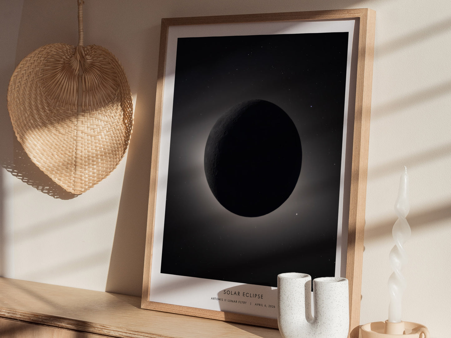 A framed poster depicting a solar eclipse, featuring a darkened moon against a soft, illuminated background. The artwork is displayed on a light wooden surface alongside a textured wall hanging.