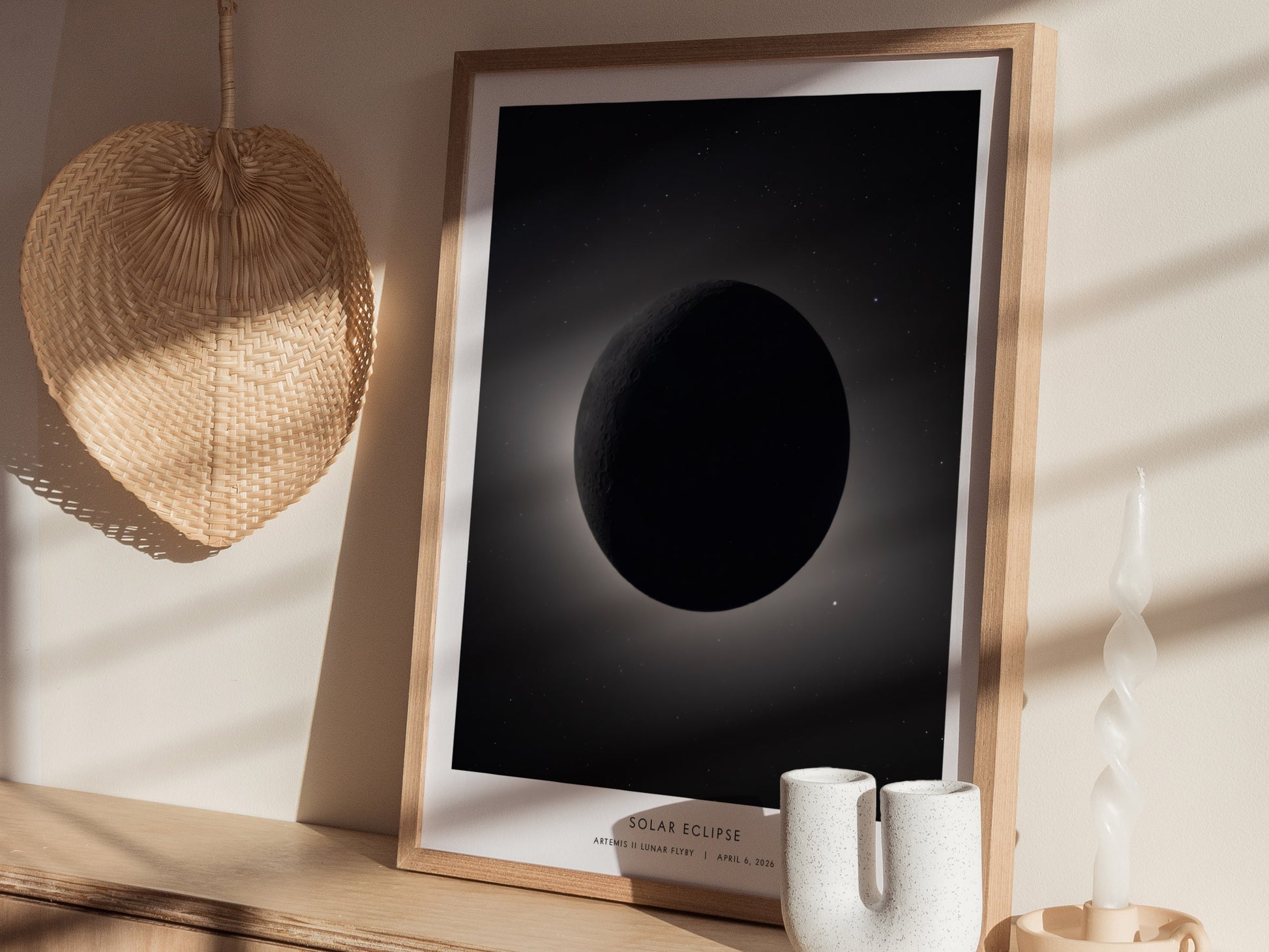 A framed poster depicting a solar eclipse, featuring a darkened moon against a soft, illuminated background. The artwork is displayed on a light wooden surface alongside a textured wall hanging.