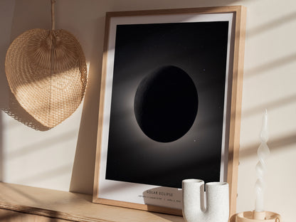 A framed poster depicting a solar eclipse, featuring a darkened moon against a soft, illuminated background. The artwork is displayed on a light wooden surface alongside a textured wall hanging.