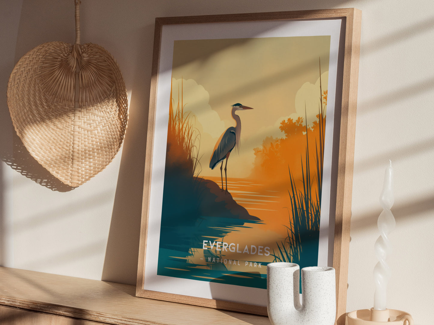 A framed minimalist poster of a heron by water at sunset, with warm orange and teal hues, is displayed on a wooden surface beside a wicker fan and a twisted.