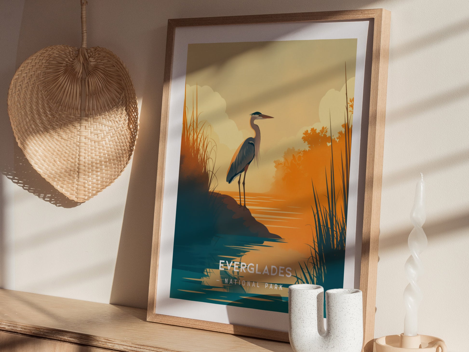 A framed minimalist poster of a heron by water at sunset, with warm orange and teal hues, is displayed on a wooden surface beside a wicker fan and a twisted.