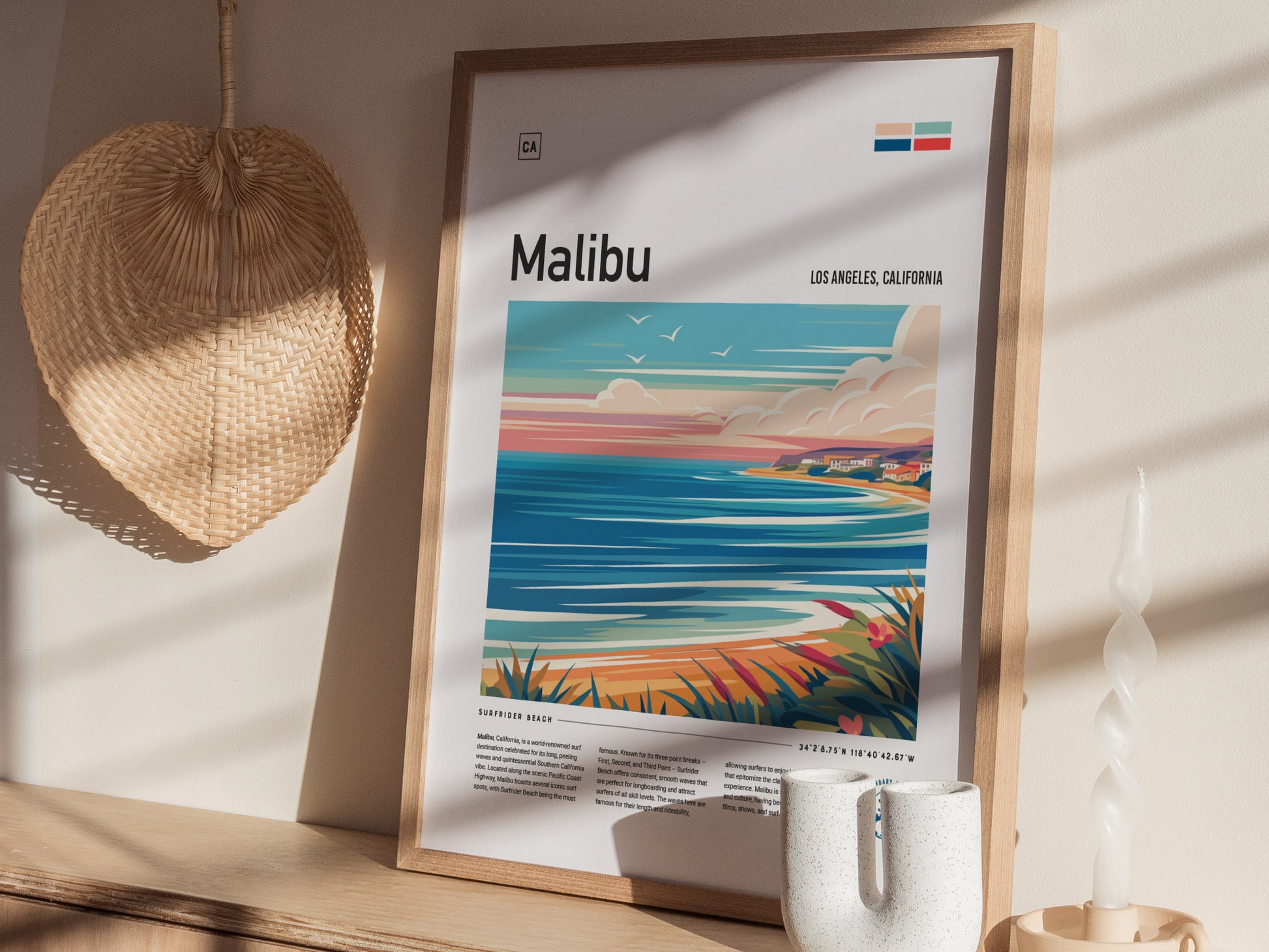 A framed Malibu poster with an abstract coastal design sits on a wooden shelf, accompanied by a woven wall decoration and a twisted white candle.
