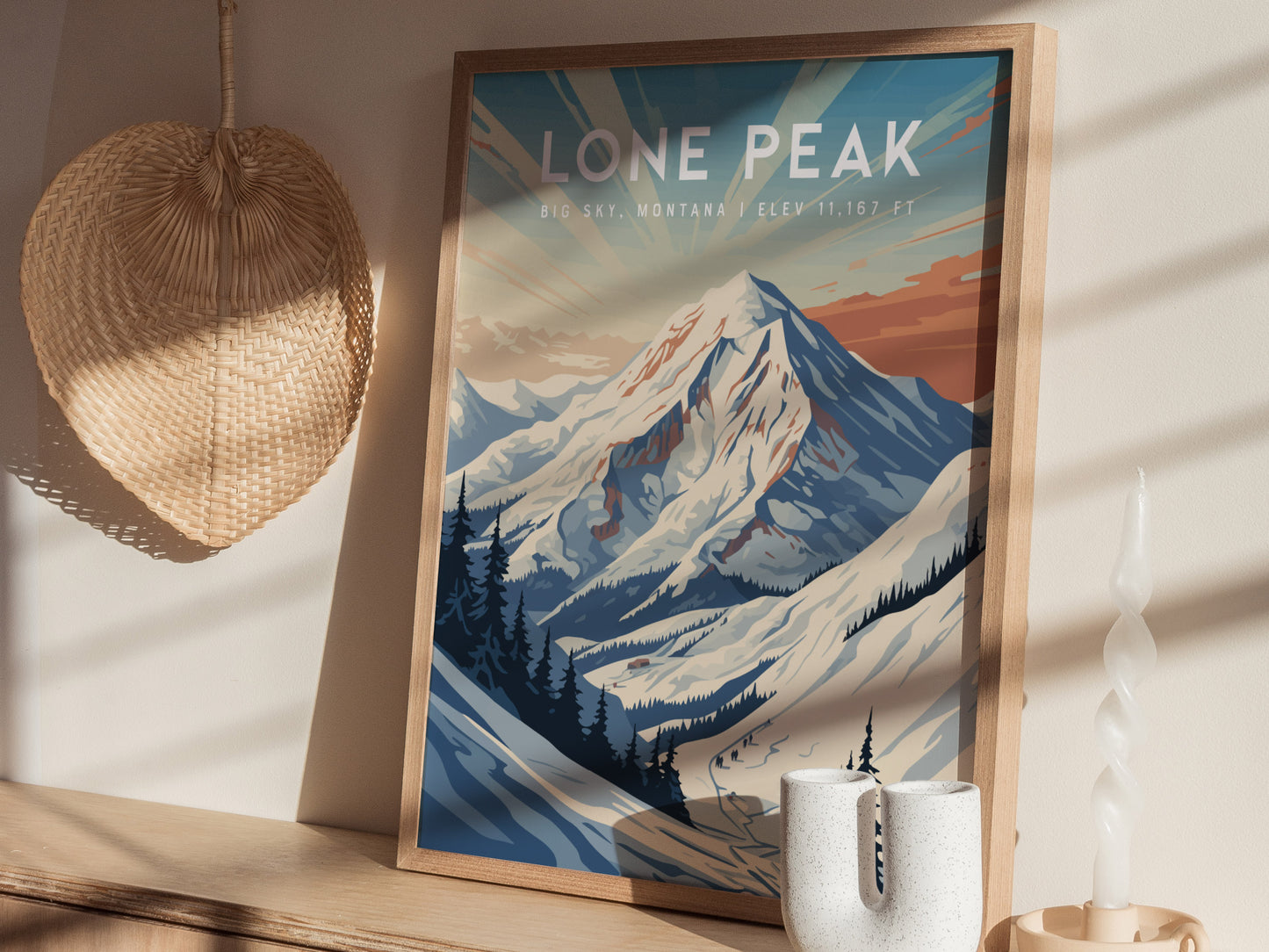 A framed poster of Lone Peak, Montana, depicts a snow-covered mountain under a colorful sky. Its displayed on a wooden surface with a wicker decoration and candle nearby.