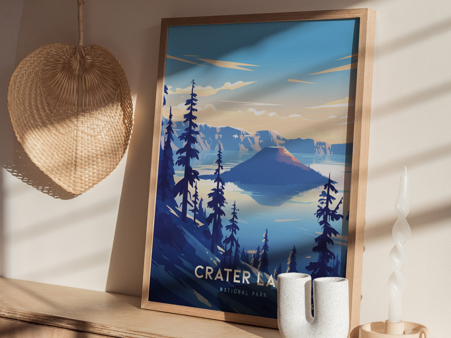 A framed poster of Crater Lake National Park features vibrant blues and a serene landscape, displayed on a wooden shelf next to a wicker basket and a white candle.