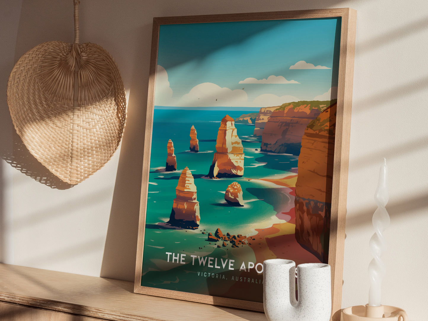 A framed poster of the Twelve Apostles in Australia is displayed on a wooden surface, with a woven basket and a twisted white candle beside it.