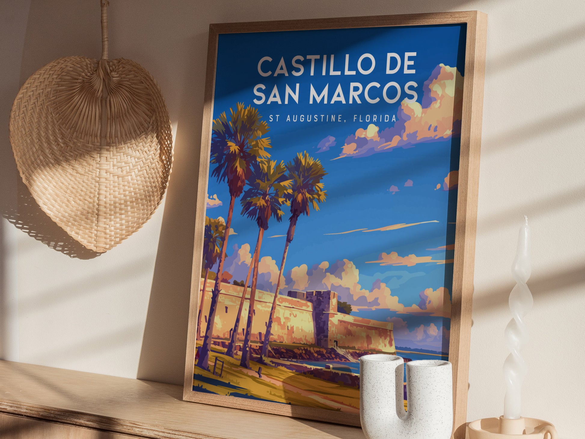 A framed poster of Castillo de San Marcos is displayed on a wooden shelf, alongside a woven fan, a white candle, and a decorative ceramic piece.