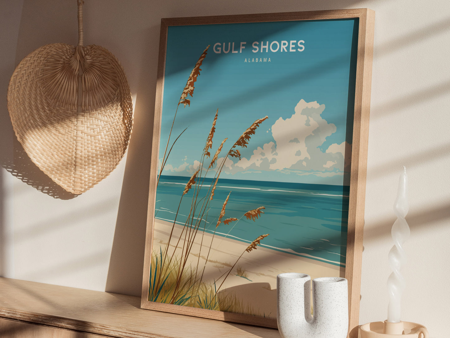 A framed Gulf Shores, Alabama poster depicting a serene beach scene with sea oats, blue waves, and fluffy clouds is displayed on a wooden shelf beside a woven fan and.