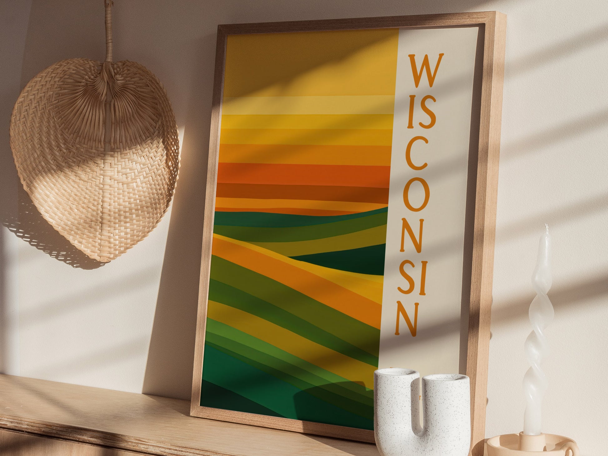 A framed abstract poster with green and gold geometric patterns features the word Wisconsin vertically. Its displayed on a wooden surface next to a wicker fan and a twisted candle.