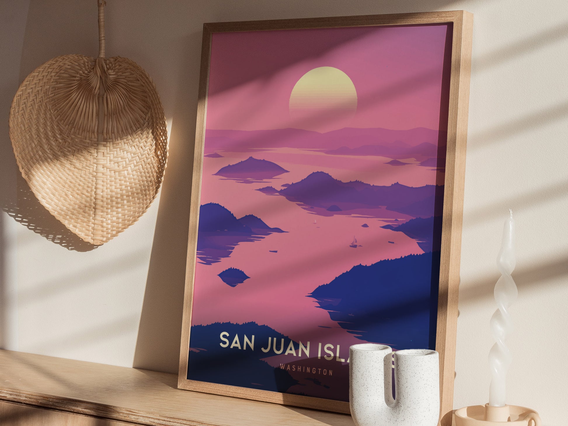 A framed San Juan Islands poster with a pink and purple sunset scene is displayed on a wooden shelf next to a wicker fan and a modern candle holder.