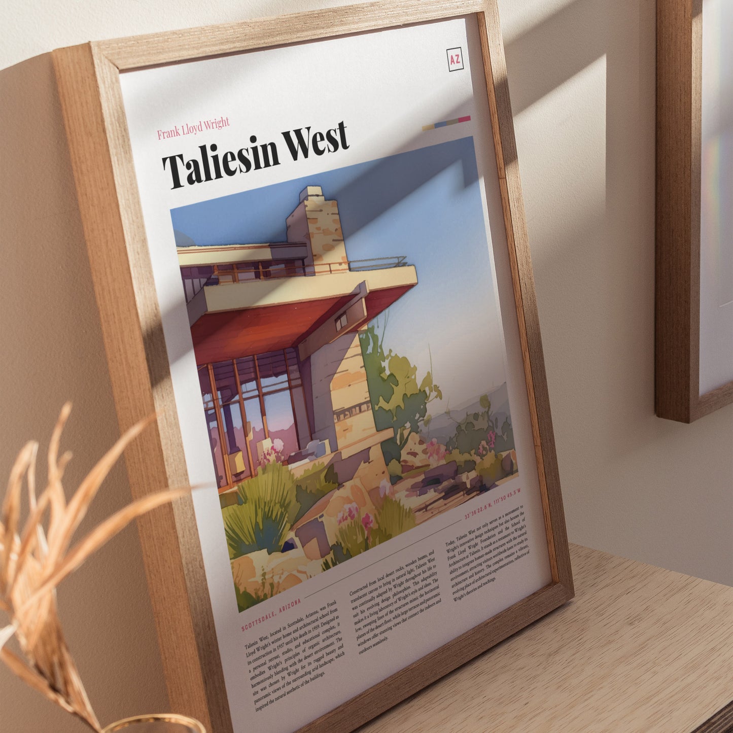A framed poster of Taliesin West is displayed on a wooden surface, showcasing Frank Lloyd Wrights architectural design with desert plants and a modern structure.