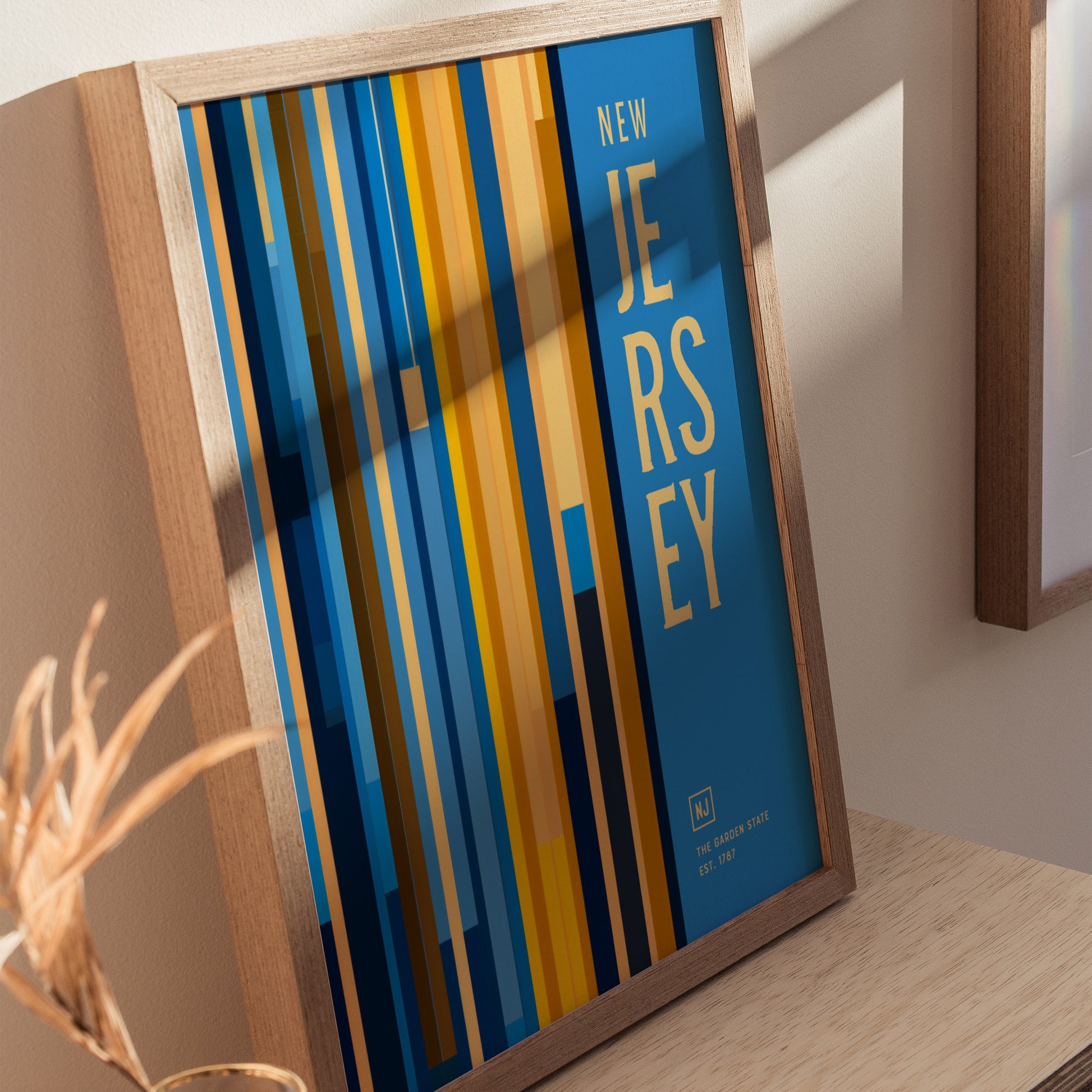 A framed New Jersey poster features vertical blue and yellow stripes with bold text. It rests on a wooden surface, casting shadows on a light-colored wall.