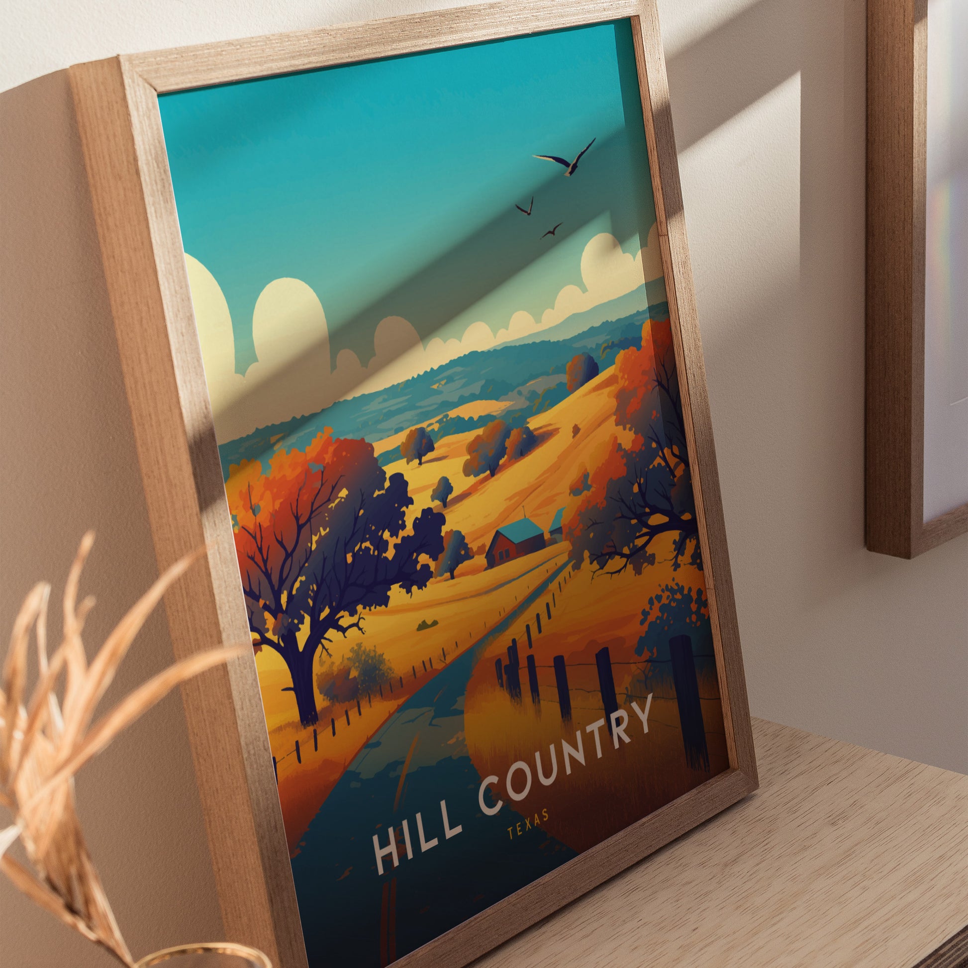 A framed poster titled Hill Country Texas features vibrant hills, trees, and a small house under a blue sky with birds flying, displayed on a wooden surface.