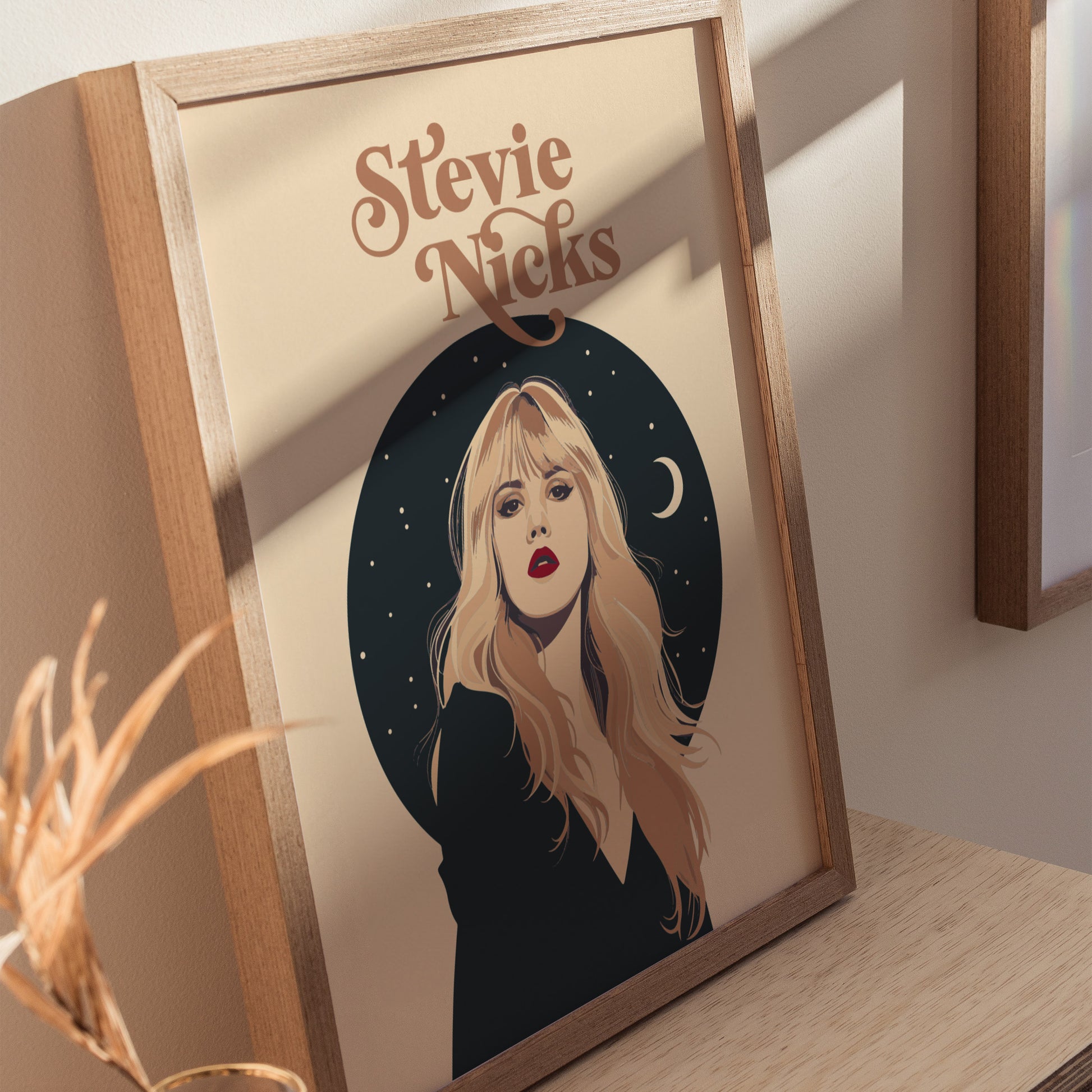 Framed poster of Stevie Nicks featuring a minimalist earth-tone portrait with celestial stars and a crescent moon, set on a wooden table with soft lighting.
