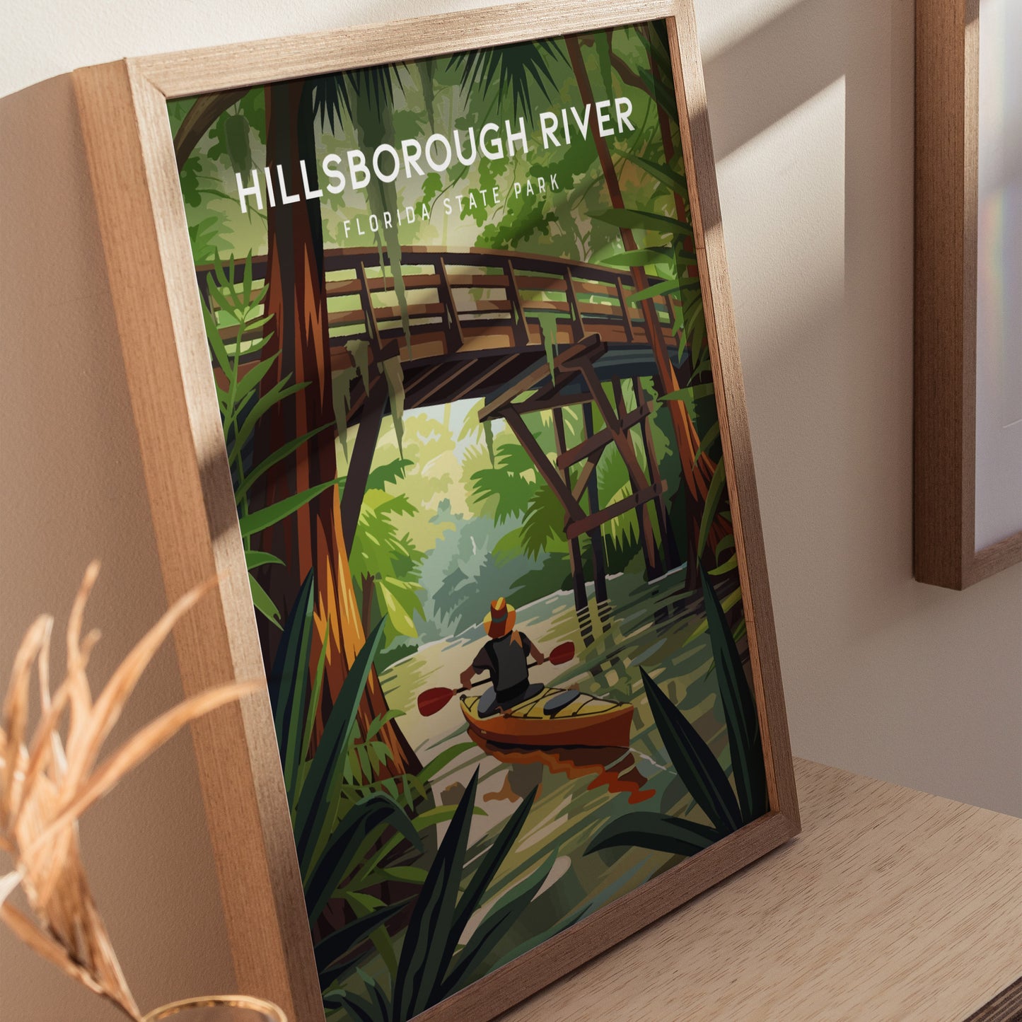 A framed poster shows a kayaker paddling on a river beneath a wooden bridge, surrounded by lush green foliage and tall trees. The text reads Hillsborough River, Florida State Park.