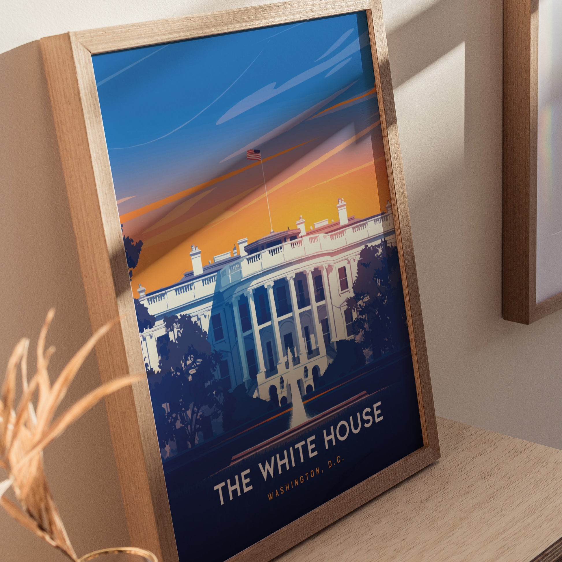 Framed poster of the White House with a vibrant sunset background, featuring bold colors and clean lines. The artwork is displayed on a wooden surface, creating a warm ambiance.