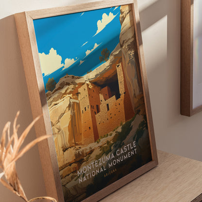 Framed poster of Montezuma Castle National Monument featuring a stylized illustration of cliff dwellings under a bright blue sky with fluffy clouds. The poster leans against a light-colored wall.