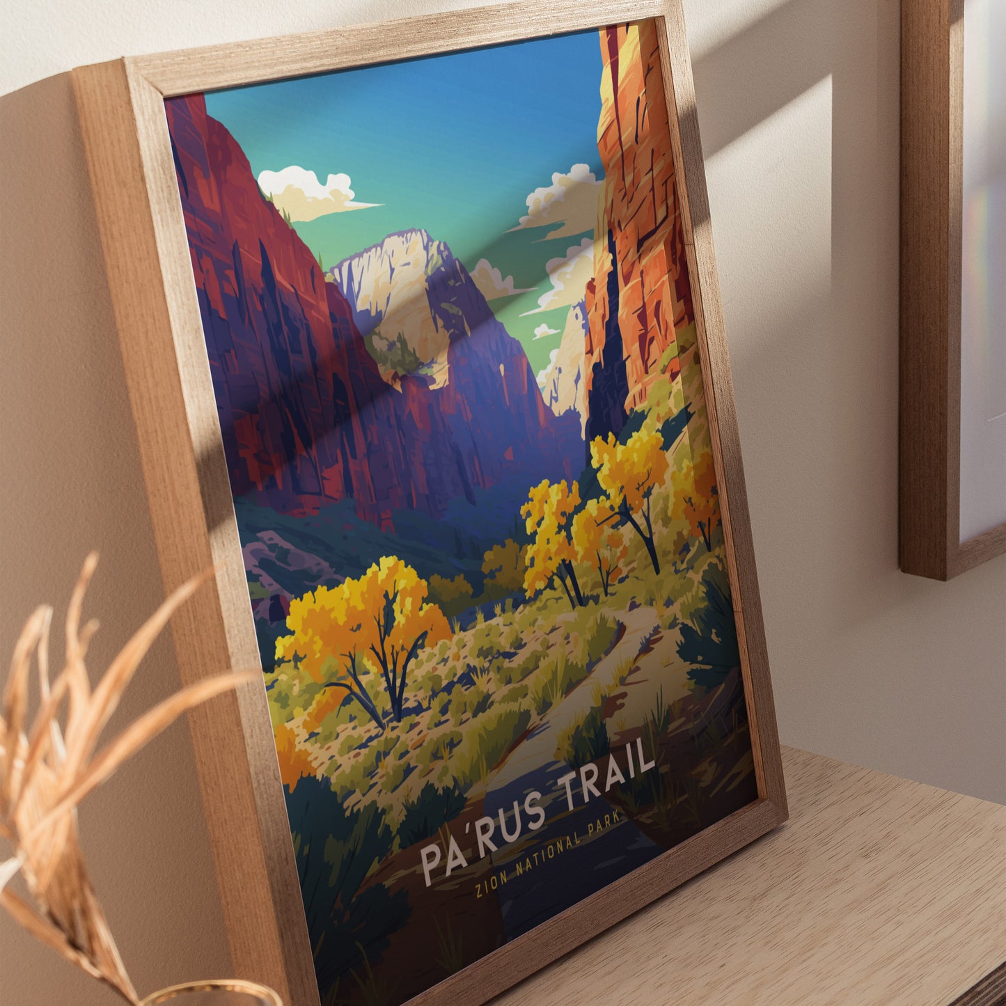 A framed poster depicting a colorful illustration of the Parus Trail in Zion National Park, with vibrant trees and dramatic canyon landscapes, rests on a wooden surface.
