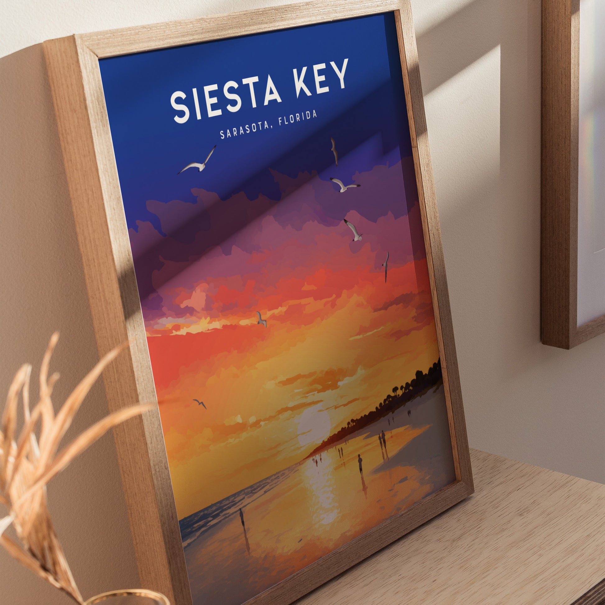 A framed poster of a vibrant sunset over Siesta Key Beach, featuring hues of orange, purple, and yellow with silhouettes of birds and people along the shoreline.