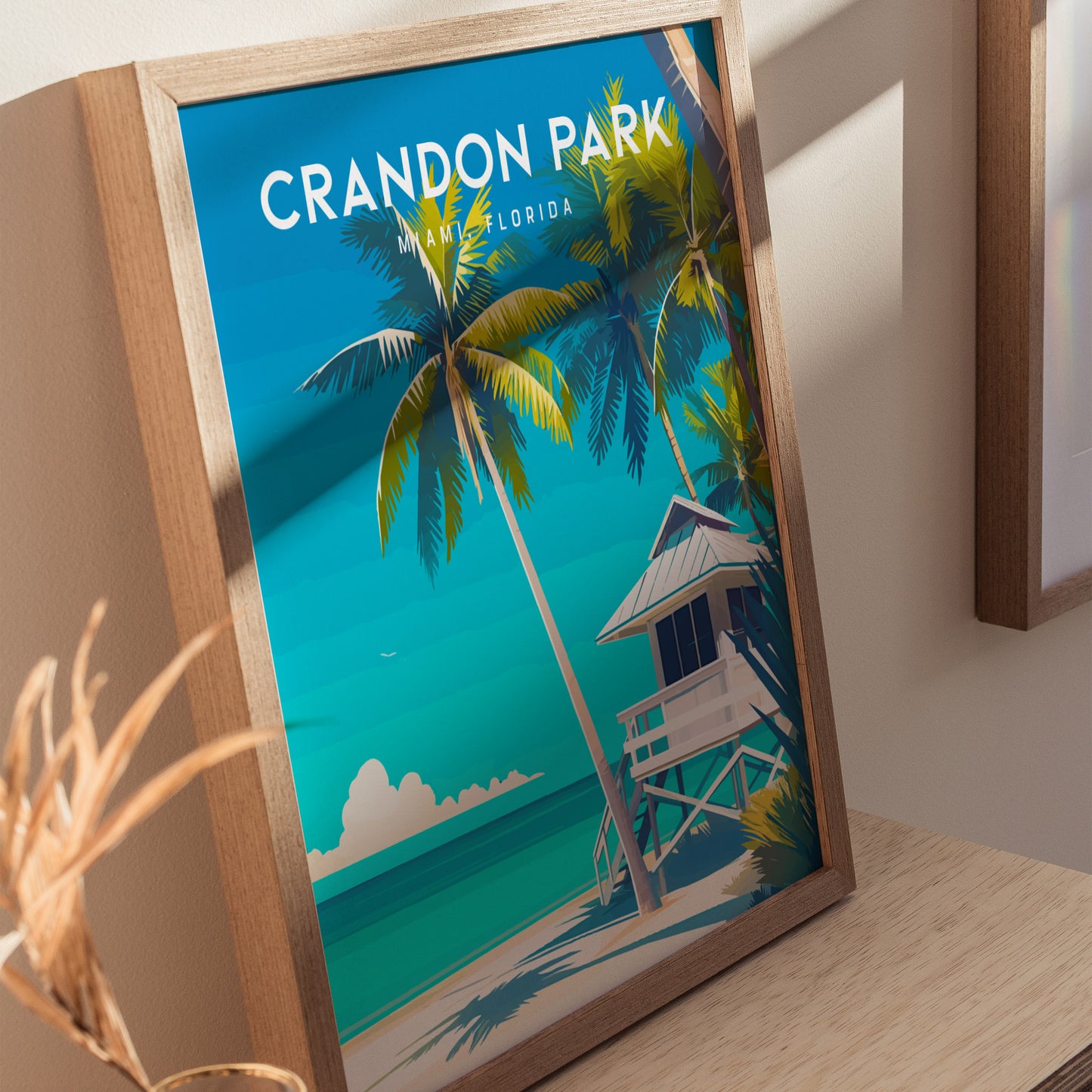 A framed poster of Crandon Park Beach, Miami, Florida, featuring vibrant palm trees, a white lifeguard tower, and turquoise waters under a clear blue sky. The artwork is displayed on a wooden surface.