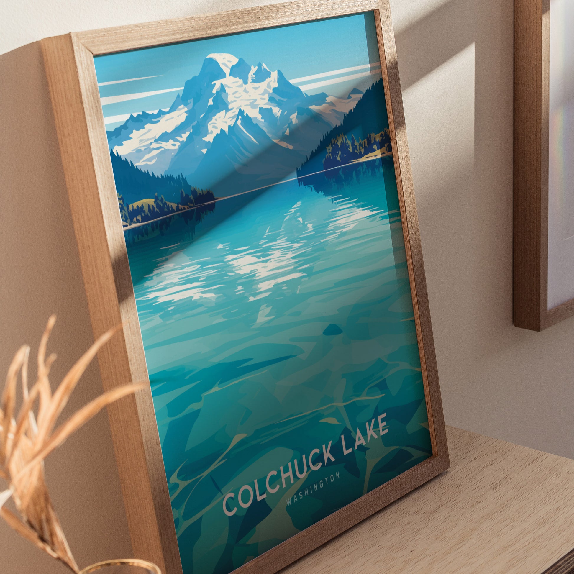 Framed artwork of Colchuck Lake with turquoise waters and snow-capped mountains under a blue sky. The poster leans against a wall on a wooden surface.
