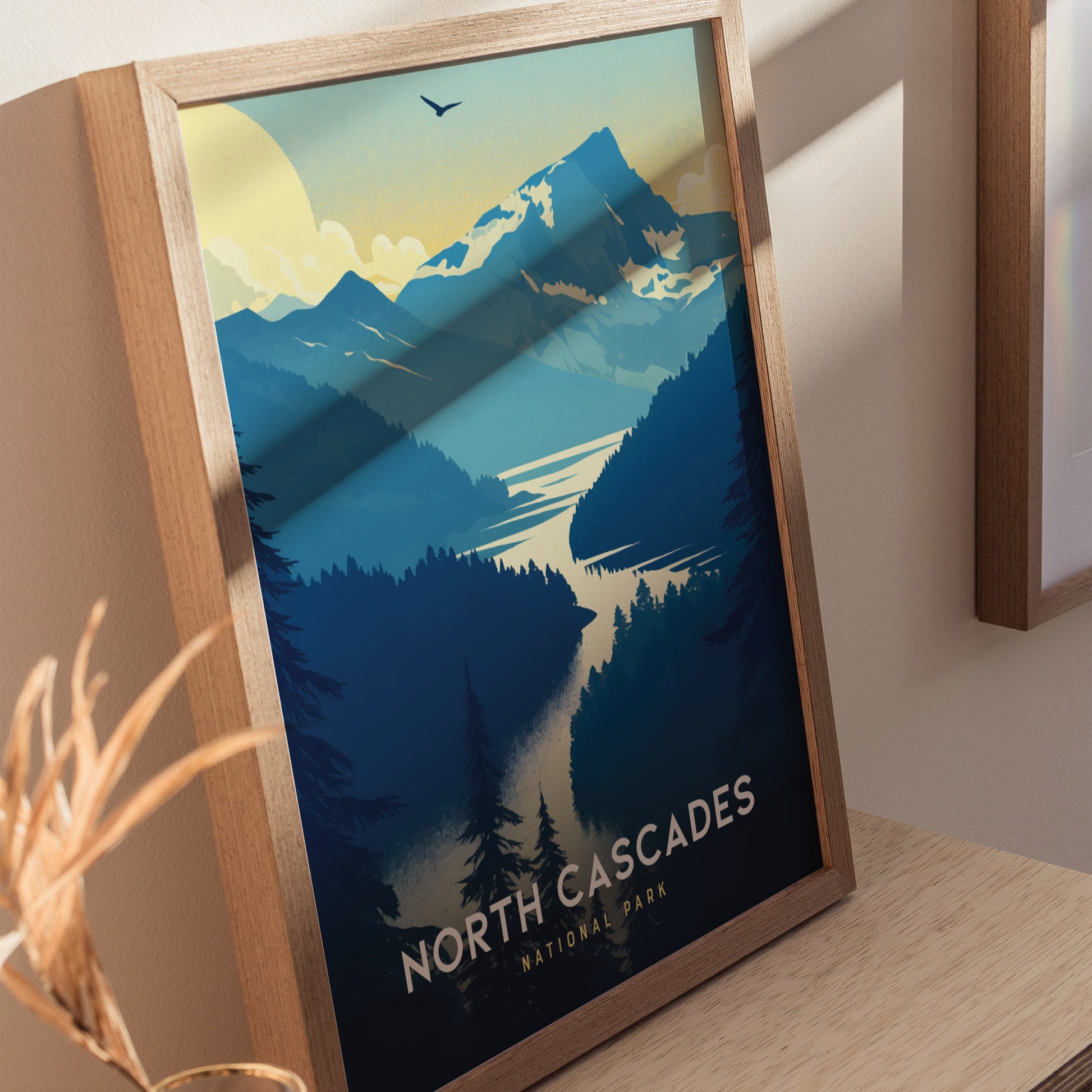 Framed poster of North Cascades National Park showcasing vibrant blue mountains, a flowing river, and a soaring bird under a bright sun. Ideal for nature-themed decor.