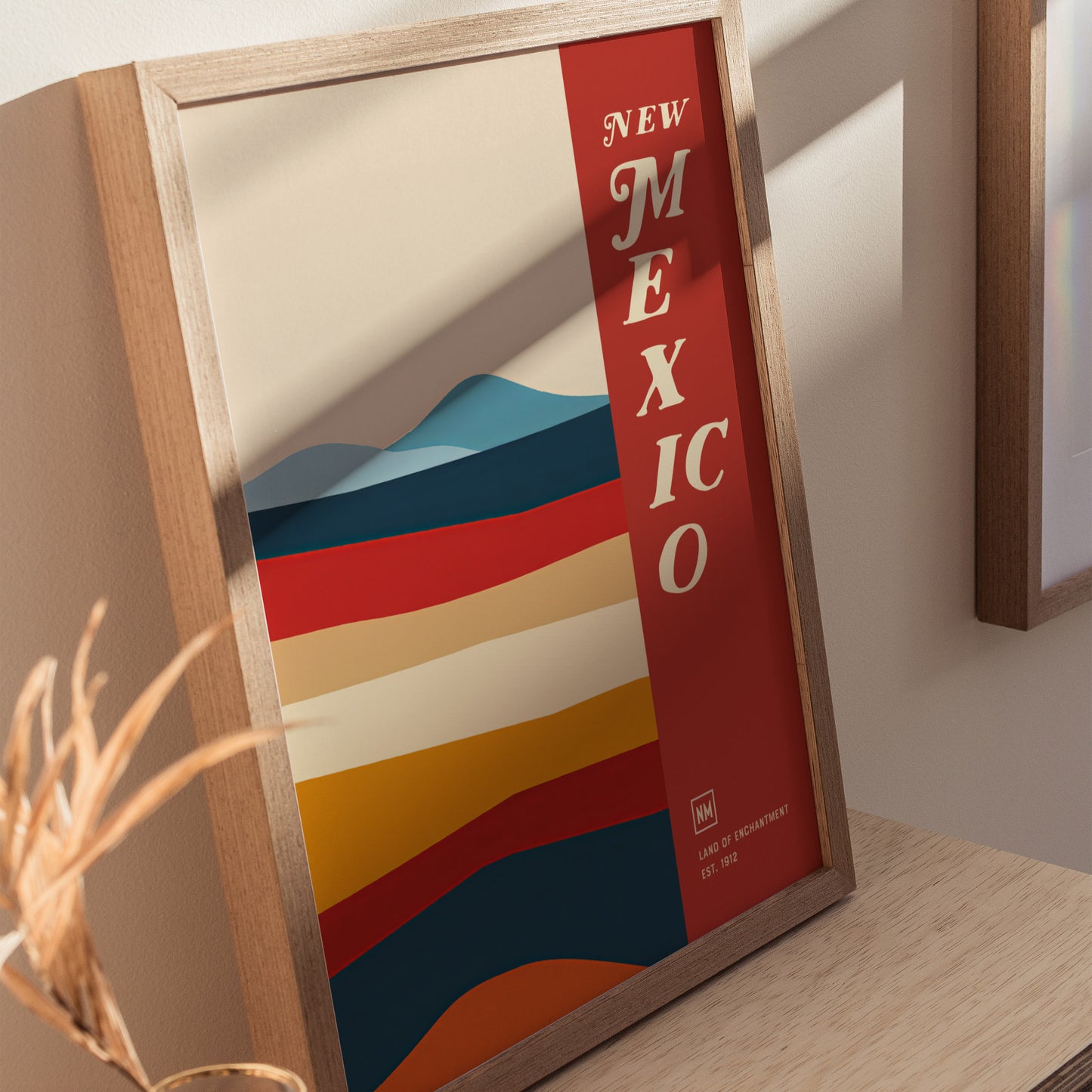 A framed poster features colorful horizontal stripes representing desert landscapes with the words New Mexico vertically on a red band. Its displayed on a wooden surface with soft lighting.