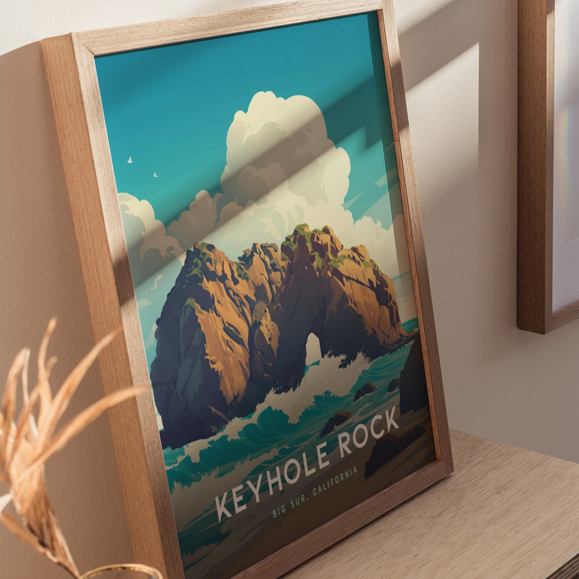 A framed poster of Keyhole Rock at Big Sur, California, is displayed on a wooden surface. The artwork features dramatic waves and a rocky arch under a bright blue sky with clouds, capturing a serene coastal scene.