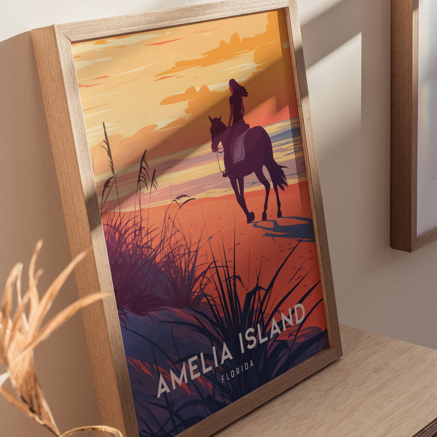 A framed poster depicting a silhouette of a woman on horseback riding along a beach at sunset, with vibrant orange and purple hues and the text Amelia Island Florida.