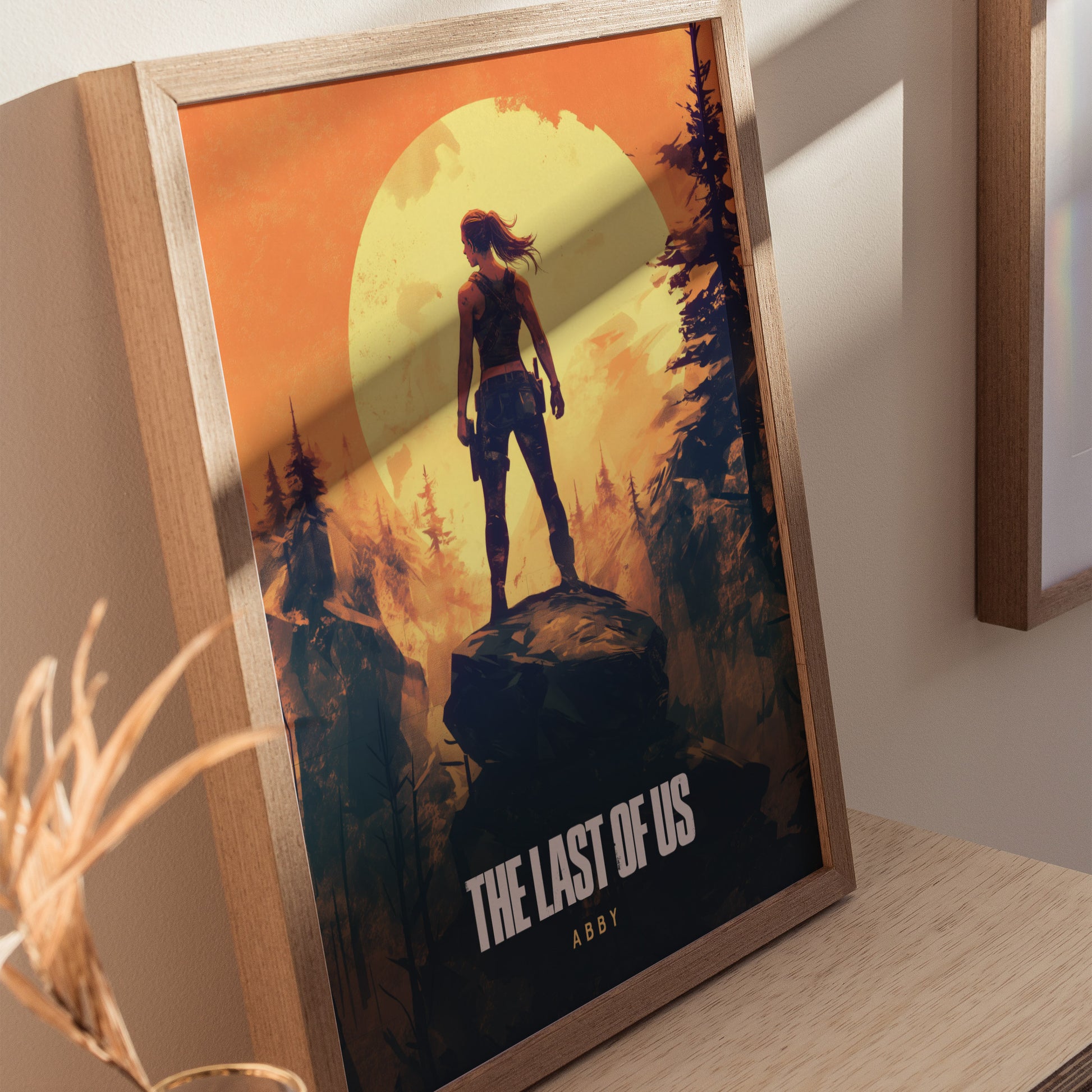Framed poster of The Last of Us featuring Abby silhouetted against a large yellow sun in a forest setting, displayed on a wooden surface.