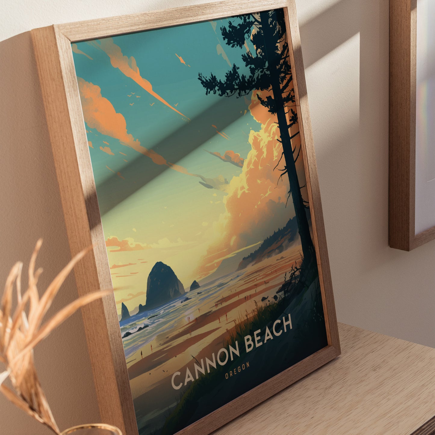 A framed poster of Cannon Beach, Oregon, features a vibrant sunset over the ocean with Haystack Rock and silhouettes of trees. It rests on a light wooden surface.