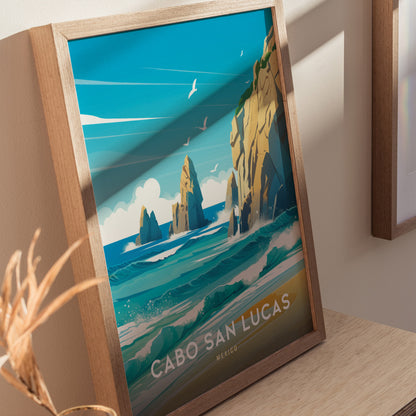 A framed poster of Cabo San Lucas features vibrant turquoise waters, rocky cliffs, and seagulls under a bright blue sky. The artwork rests on a wooden surface.