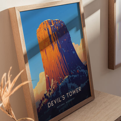 A framed poster of Devils Tower, featuring a vibrant illustration of the geological formation against a blue sky, is displayed on a wooden surface.