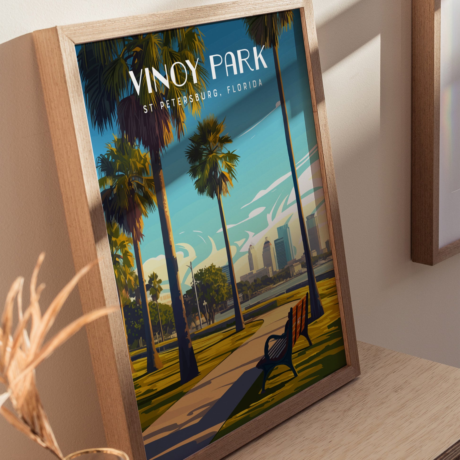 Framed Vinoy Park poster featuring palm trees, a park pathway, and a view of the St. Petersburg skyline, displayed on a wooden surface. Bright, scenic artwork captures Floridas beauty.