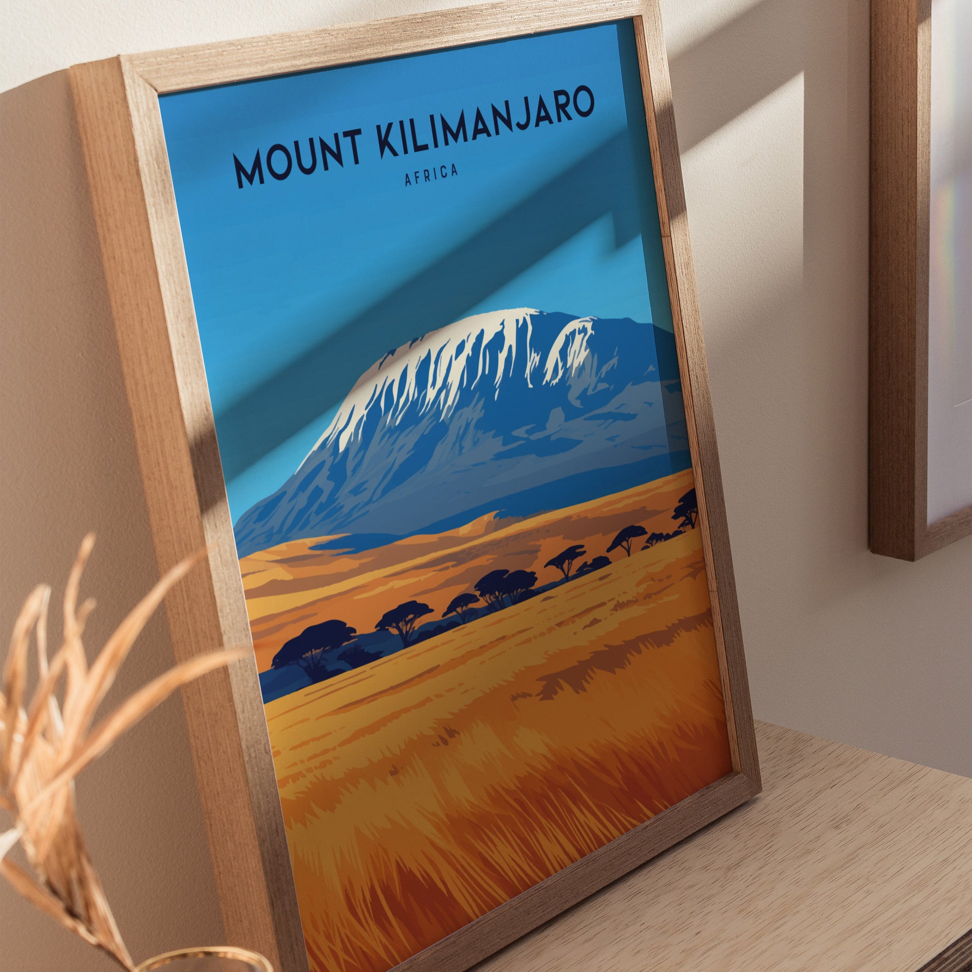 Framed poster of Mount Kilimanjaro with a snow-capped peak, set against a blue sky and golden savanna, displayed on a wooden surface. Shadows and lighting create a warm ambiance.