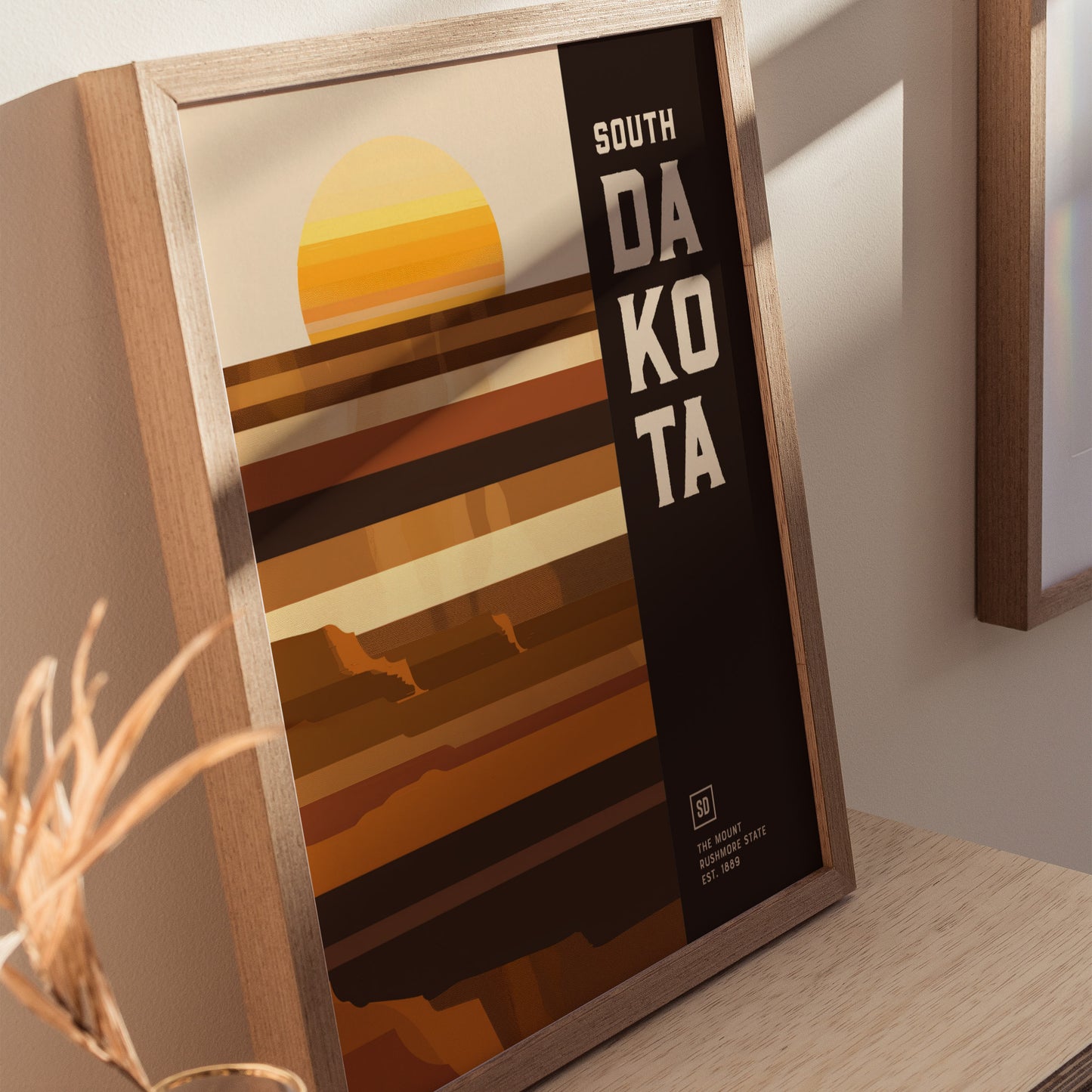 Framed poster with geometric design features a stylized sunset and earthy tones, highlighting South Dakota in bold text. It rests on a light wooden surface.