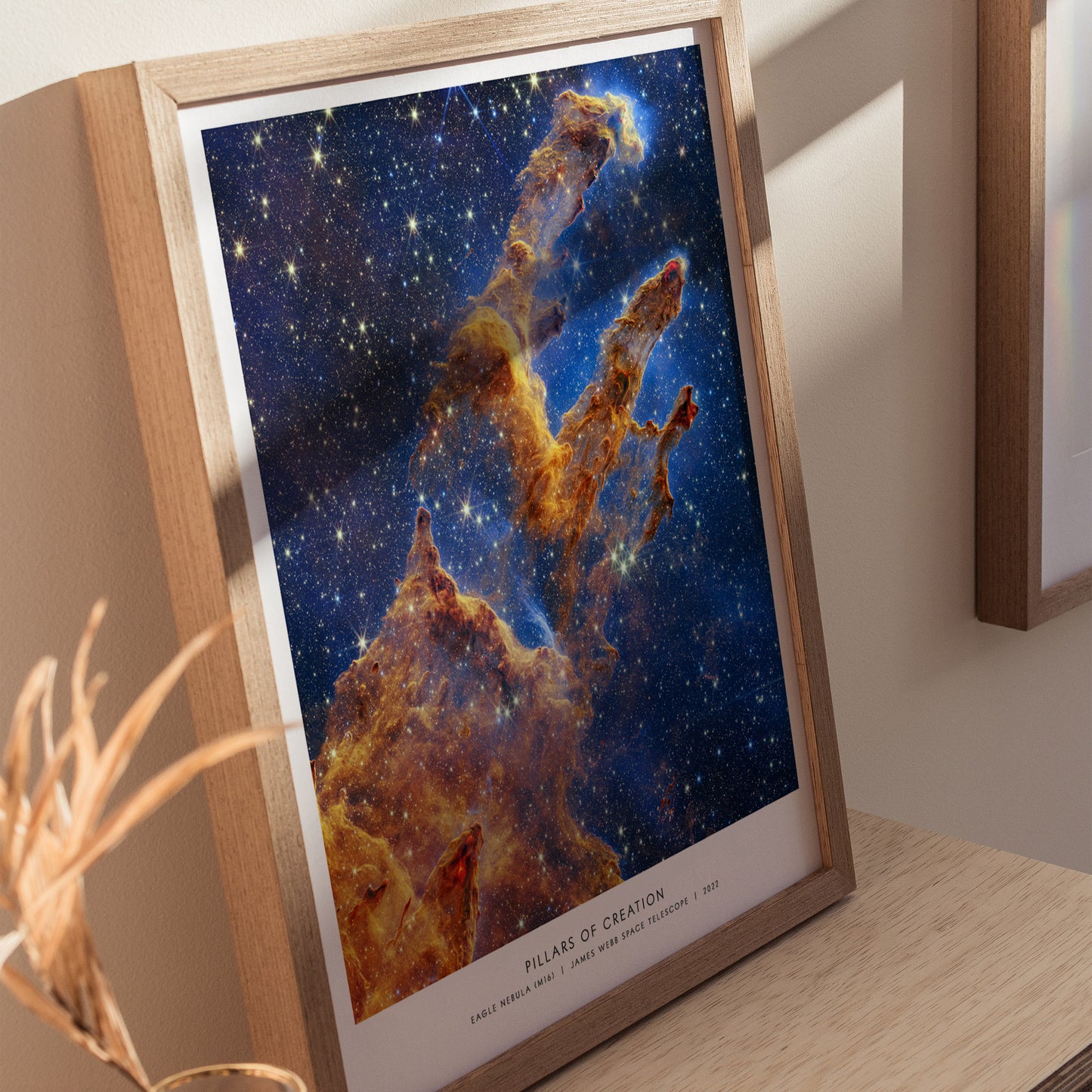 Wood frame version of the Pillars of Creation James Webb Space Telescope poster leaning against a wall, featuring bright star fields and glowing nebula clouds.