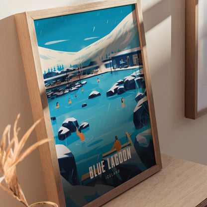 Framed poster depicting Blue Lagoon in Iceland, featuring people swimming in milky blue water surrounded by black lava rocks and snowy mountains. Displayed on a wooden tabletop.