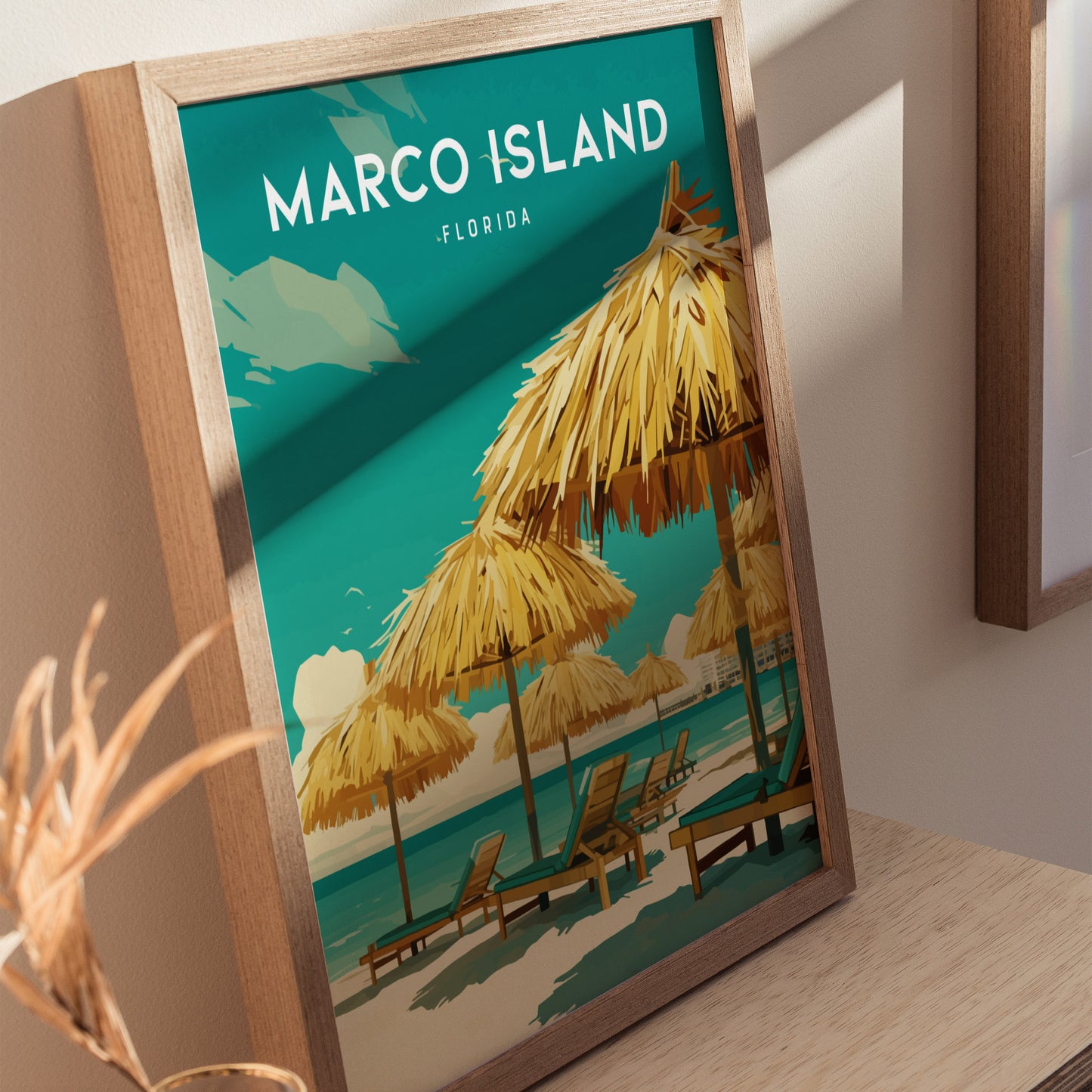A framed poster featuring Marco Island Florida with illustrations of beach umbrellas and lounge chairs on a sandy shore against a turquoise sea, displayed on a wooden surface.