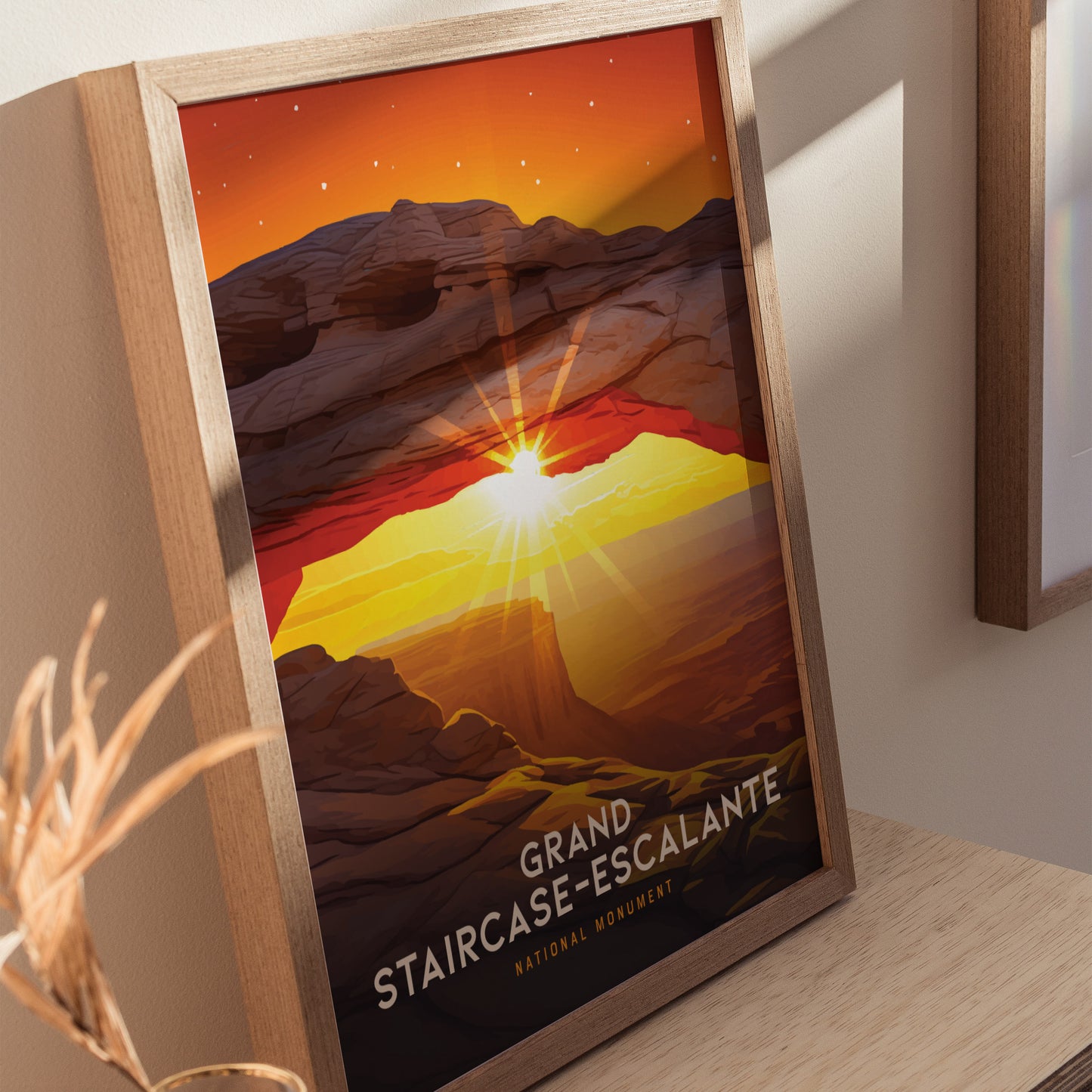 A framed poster of Grand Staircase–Escalante National Monument depicts a vibrant sunset arch with a glowing sun and colorful sky. The artwork is placed on a wooden surface.