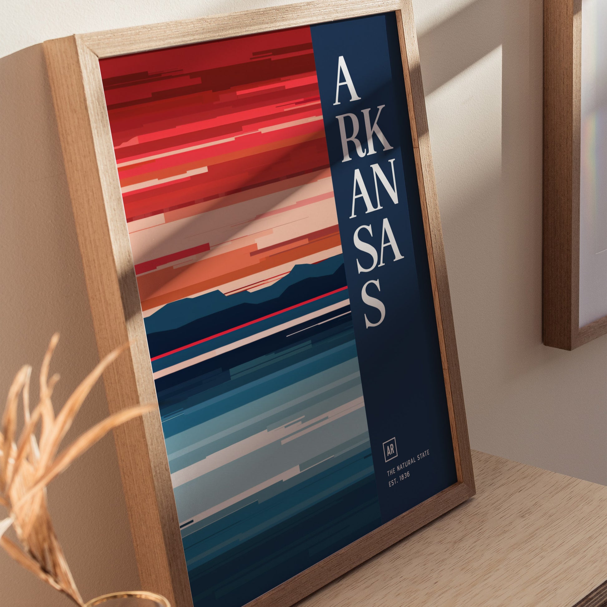 Framed Arkansas poster with abstract horizontal stripes in vibrant red, orange, and blue tones, featuring bold vertical text. Displayed on a wooden surface with natural light.