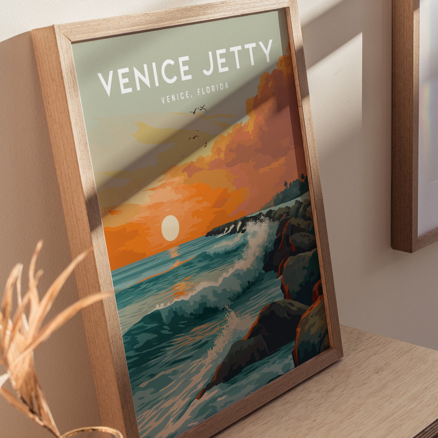 A framed poster titled Venice Jetty shows a vibrant sunset over the ocean, with waves crashing against a rocky jetty. The artwork features warm orange.